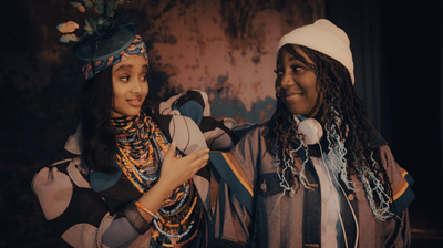 Two young people looking at each other, one with light brown skin wearing colorful jewelry and clothing, and one with brown skin wearing a beanie hat and headphones around their neck.