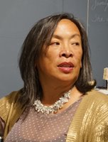 Cynthia is looking off to the right side, with a blackboard in the background. She has light brown skin and dark brown, straight hair falling to her shoulders, and is wearing pink lipstick, a white fuzzy-textured necklace, a lavender colored top with white polka dots, and a metallic gold cardigan.