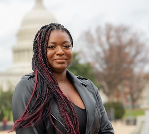 Quanice is standing outdoors, wearing a black leather jacket and a black top. She has warm brown skin and her hair is in long, black and red braids. The U.S. Capitol Building and some trees are out of focus in the background.