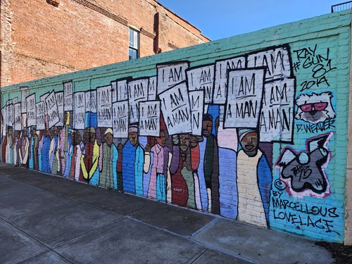 Marcellous Lovelace's mural entitled I Am A Man. It depicts a crowd of people with brown skin, wearing colorful clothing and holding up signs that say "I Am A Man", over a turquoise colored background.